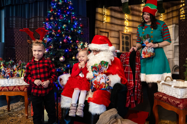 Dwójka dzieci pozuje do zdjęcia ze świętym Mikołajem. W tle świąteczne dekoracje i kobieta przebrana za elfa. 